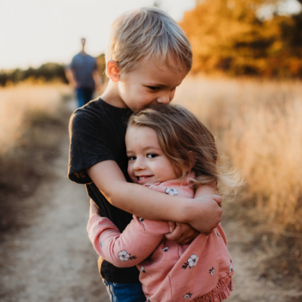 Young brother and sister hug in open field with dad at a distance