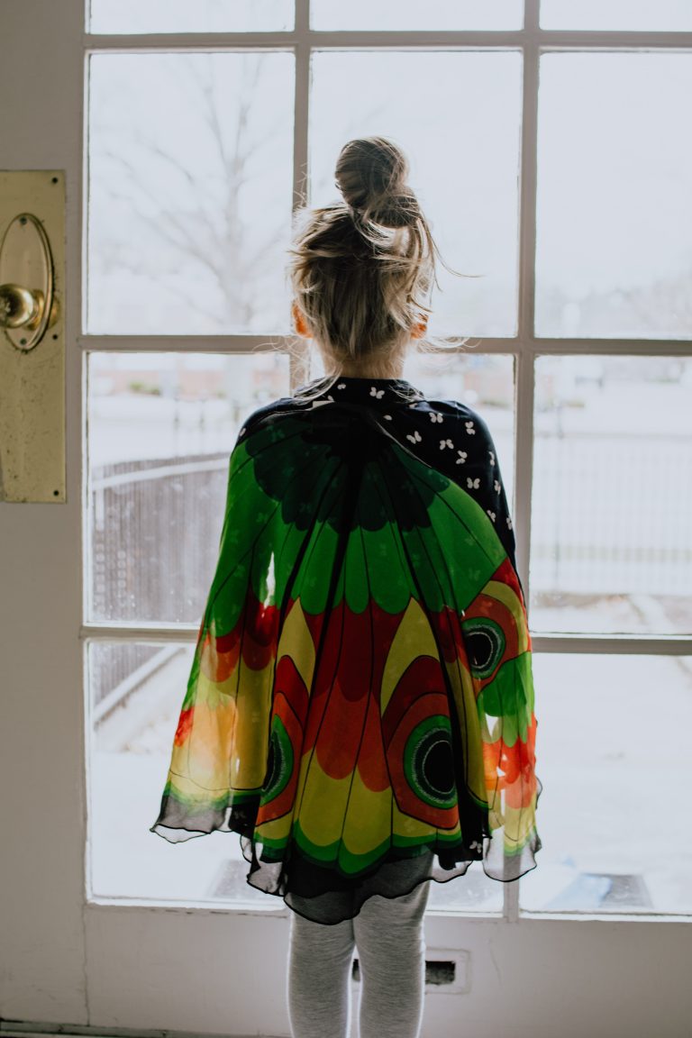 Child stares out a glass door wearing multi coloured costume