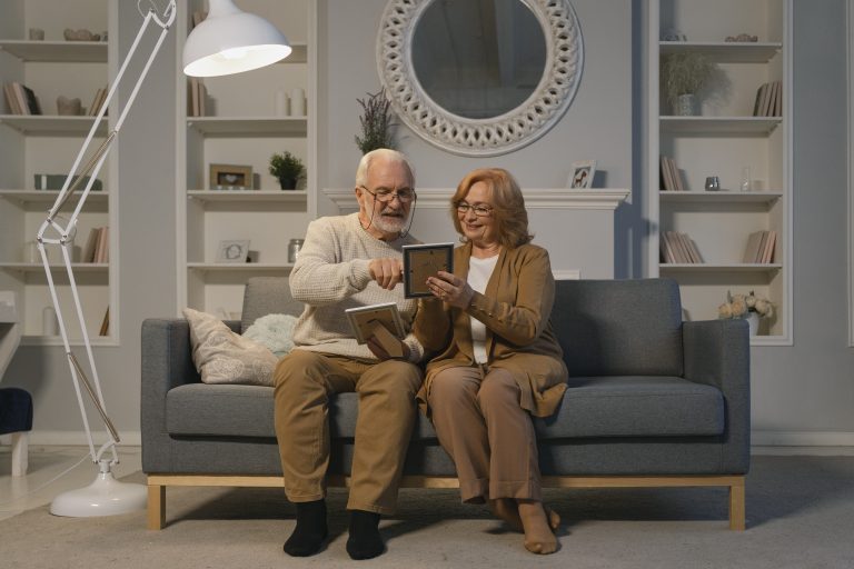 Elderly couple sit on sofa looking at picture frames smiling