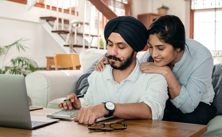 Sikh couple sit at dinging table looking through laptop smiling