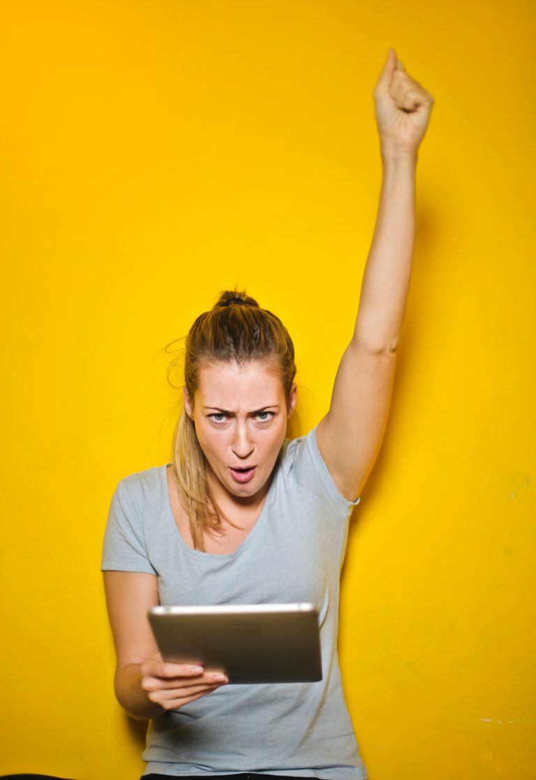 Excited woman raises a fist up as she reads a tablet standing in front of an orange background