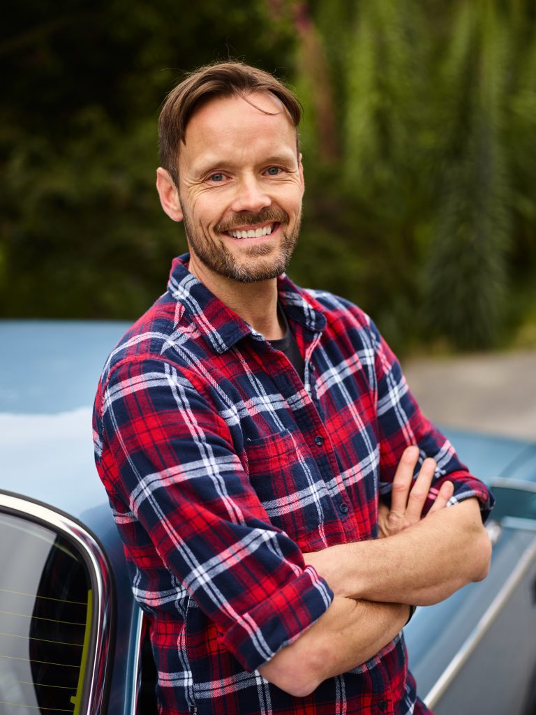Man smiles with arms crossed standing leaning on blue car