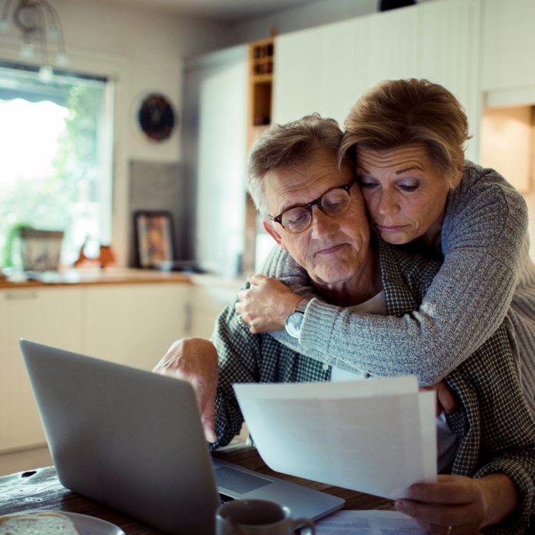 Wife hugs husband from the back as they look over bills on paper and a laptop