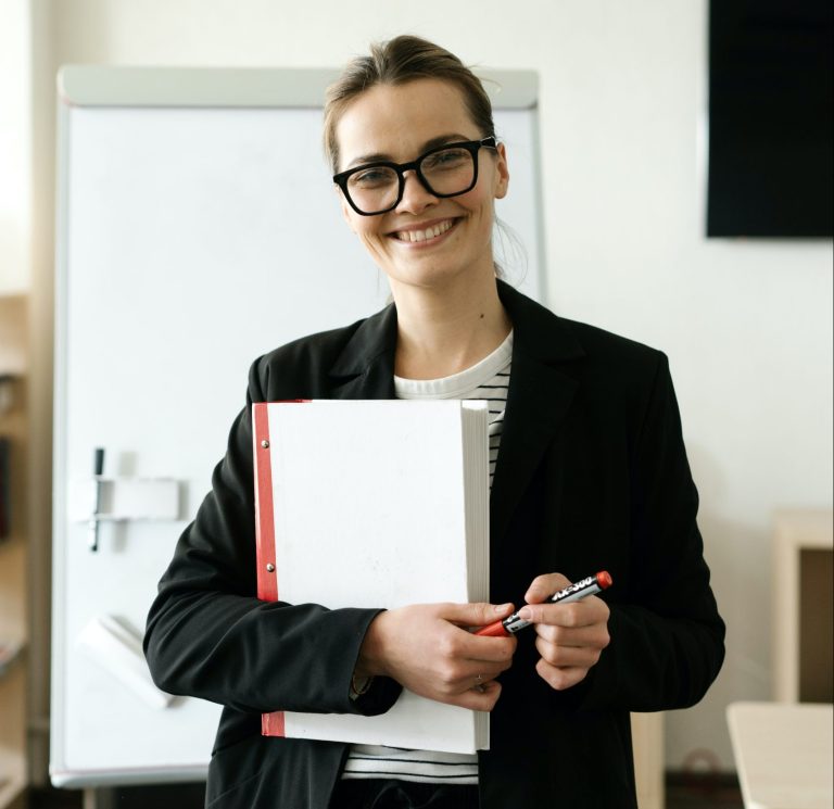 Woman wearing glasses smiles in the office while holding binder and markers