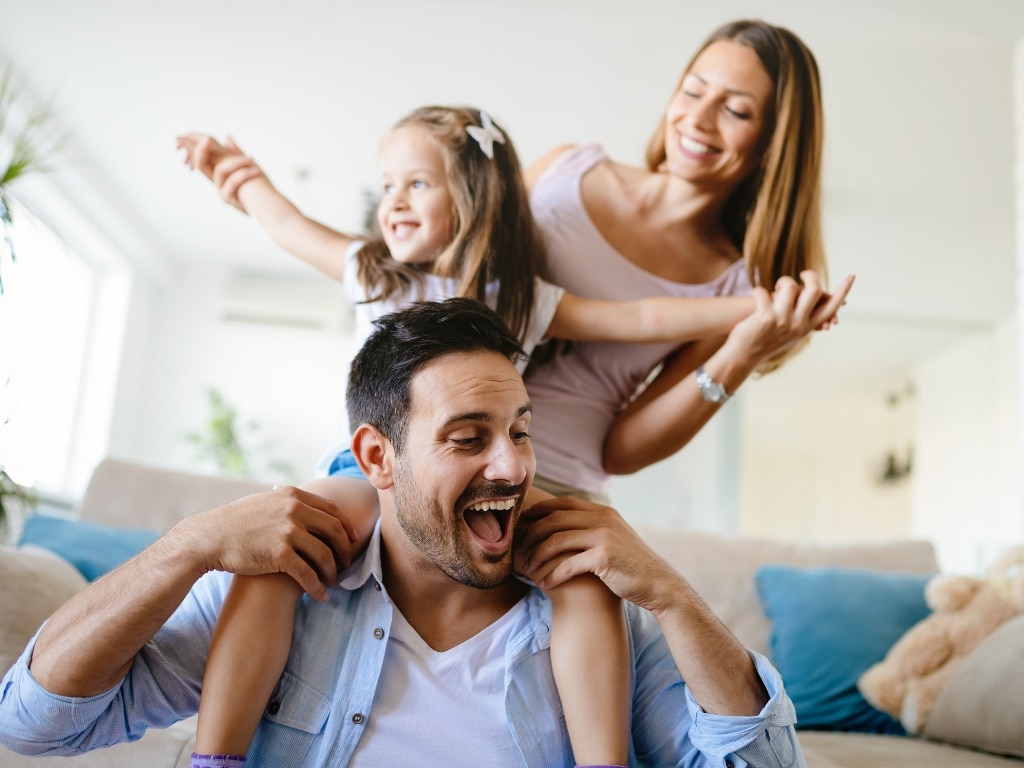 Father laughing with his little girl on his shoulders while the wife holds the daughter's hands, all looking happy and content in the living room, highlighting Scullion LAW's commitment to family well-being.