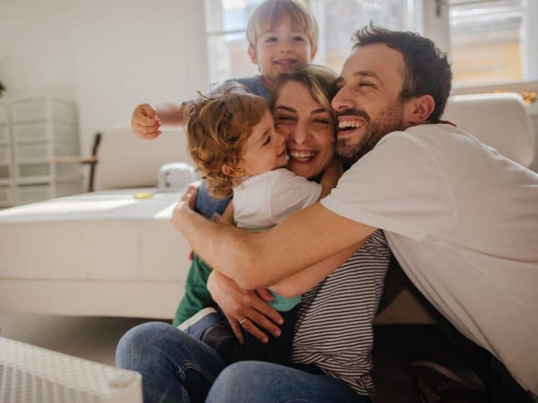 A mother and father share a joyful group hug with their two young children in their living room. Everyone is smiling and laughing, creating a warm and happy family moment.