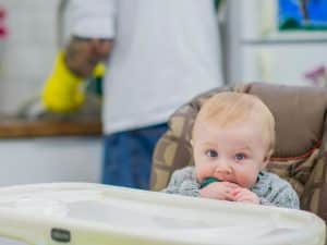 An infant with bright blue eyes sits in a high chair, sucking on a teether, while a parent does the dishes in the background.