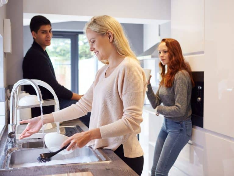 Blonde woman washing dishes at the kitchen sink, laughing with two housemates. They purchased the property together and have a cohabitation agreement in place.