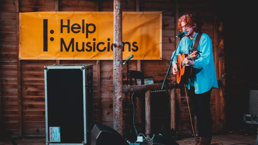 A musician plays music on a stage in front of a Help Musicians banner